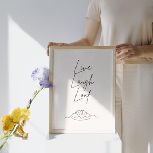 May include: A framed print with the words "Live Laugh Loaf" in elegant script, accompanied by a line drawing of a loaf of bread. The frame is light-colored, and the background is white. A sprig of yellow and purple flowers is in the foreground.