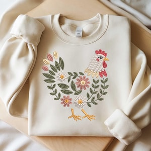 May include: Cream-coloured sweatshirt with an embroidered floral hen design. The hen is made of pink, white, and green flowers and leaves. The hen's head has a red comb and yellow beak. The sweatshirt has long sleeves with ribbed cuffs.
