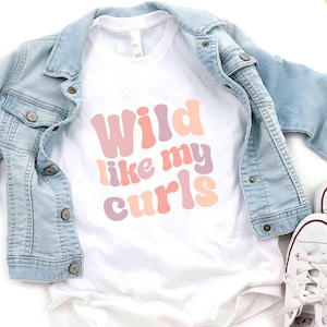 May include: A white t-shirt with the text "Wild like my curls" in pink, peach, and purple. A light blue denim jacket is draped over the shirt. White sneakers with red trim are next to the shirt.