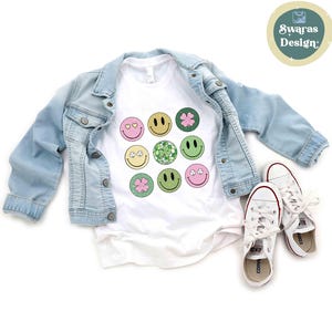 May include: White t-shirt with a colorful smiley face graphic, paired with a light blue denim jacket and white sneakers. The graphic features various smiley faces with hearts, clovers, and floral designs.