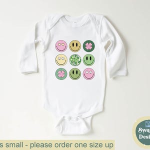 May include: White baby bodysuit with long sleeves, featuring a grid of colorful smiley faces. The smiley faces have various designs, including hearts, clovers, and disco balls. The text "Runs small - please order one size up" is at the bottom.