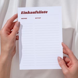 Peut inclure: Un bloc-notes blanc avec le texte allemand "Einkaufsliste" en bordeaux, tenu par deux mains. Le bloc-notes comporte des sections lignées pour l'écriture. Le fond est un tissu blanc.