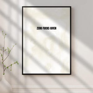 May include: A framed print with the text "ZERO FUCKS GIVEN" in black, centered on a light-colored background. The frame is black, and the print is displayed on a white wall. A white vase with a green plant is in the lower left corner.