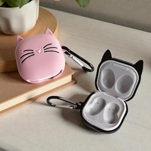 Può includere: Custodia per auricolari a forma di gatto rosa con baffi e orecchie neri, accanto a una custodia per auricolari in bianco e nero con orecchie di gatto. Entrambe le custodie hanno un moschettone nero. Su una superficie chiara.