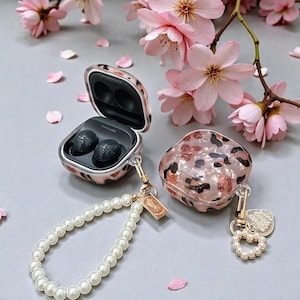 Peut inclure: Étuis pour écouteurs avec un motif léopard rose et marron, accompagnés d'une dragonne en perles et de détails dorés. Un étui ouvert révèle des écouteurs noirs. Des fleurs et pétales roses ornent l'arrière-plan.