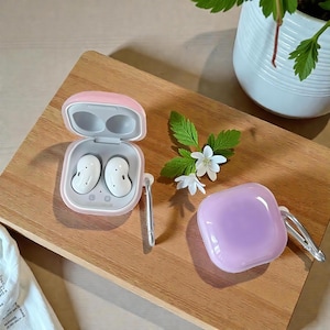 Może przedstawiać: Białe bezprzewodowe słuchawki w różowym etui, obok lawendowego etui ze srebrnym karabińczykiem. Otwarte etui odsłania słuchawki. Na drewnianej tacy, z białymi kwiatami i zielonymi liśćmi.