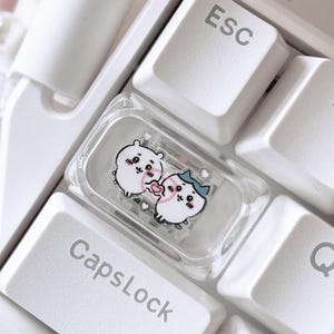 May include: A white mechanical keyboard featuring a custom transparent keycap placed between the Esc and CapsLock keys. The clear keycap displays a graphic of two small white cartoon characters holding a pink heart, set against a white keyboard base.