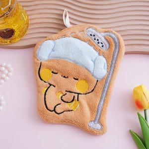 May include: A plush, tan-colored washcloth with an embroidered cartoon character design. The character is wearing a light blue hat and holding a yellow bottle. A silver shower head is also embroidered on the washcloth. A white loop is attached for hanging.