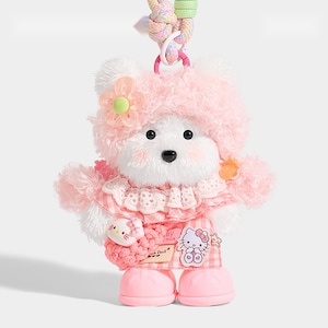 May include: A plush white bear doll wearing a pink fuzzy hat with a flower, a pink gingham dress with a lace collar, and pink boots. The doll features a Hello Kitty patch, a small Hello Kitty charm, and a pastel braided cord for hanging.