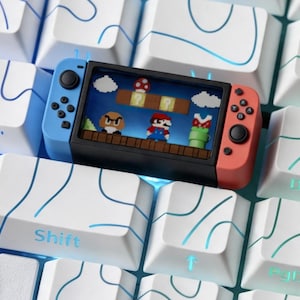 May include: A miniature handheld gaming console with a blue left controller and a red right controller, displaying a pixelated video game scene with a character, mushroom, and pipe, resting on a white computer keyboard with blue line patterns.