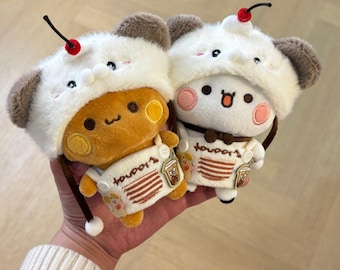 May include: Two plush toys wearing white fuzzy hoods with a red cherry on top. One toy is golden brown, the other is white. Both wear white overalls with a brown striped pocket and a small embroidered bubble tea graphic on the front.