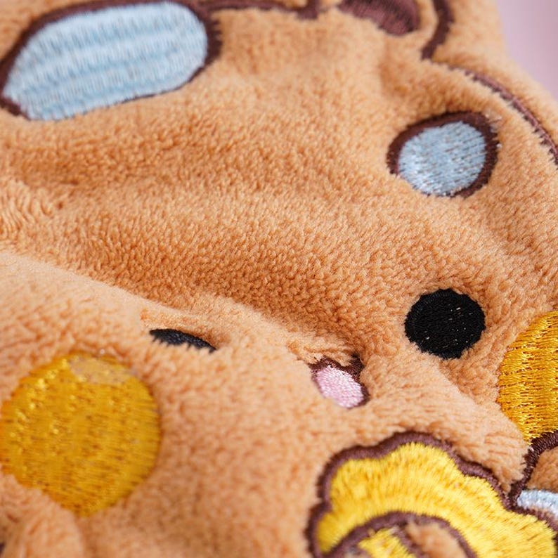 May include: Close-up of a plush, brown fabric with embroidered details. The design features a cartoon-like character with light blue, black, and yellow accents. The texture appears soft and fuzzy, suggesting a children's item.