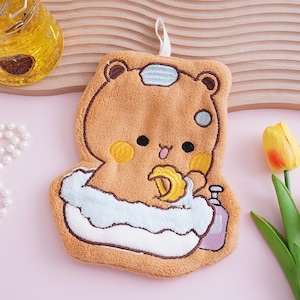 May include: A brown bear-shaped washcloth with a cartoon design, featuring a bear in a bathtub with a white fluffy border. The washcloth has a loop for hanging and a small bottle of soap.