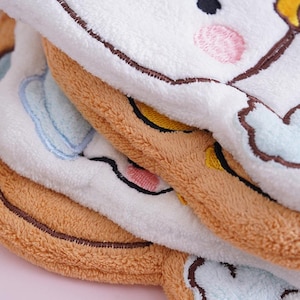 May include: Close-up of three plush, embroidered animal face towels. The towels are white, brown, and tan, with embroidered details in black, pink, and yellow. The towels are soft and fluffy, with a cartoon-like design.