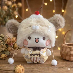 Peut inclure: Une peluche représentant un personnage blanc coiffé d'un chapeau crème duveteux surmonté d'une cerise rouge. Le personnage porte une salopette jaune et un nœud papillon marron. Il a de grands yeux ronds et des joues roses.