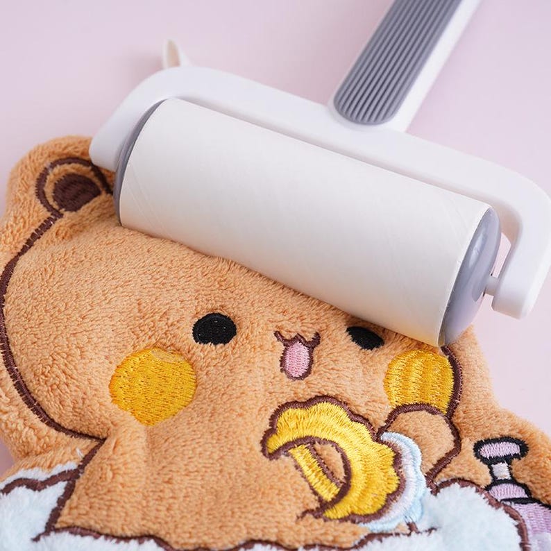 May include: A white lint roller with a gray handle is used on a brown plush towel. The towel features an embroidered cartoon bear design with yellow, black, and pink accents. The background is a soft pink.