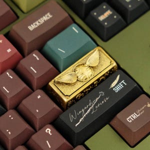 May include: A mechanical keyboard with dark brown, green, and black keycaps. One gold-coloured metal keycap features a winged sphere design. A black shift key displays the white text Wingardium Leviosa with a feather icon and a wand graphic.