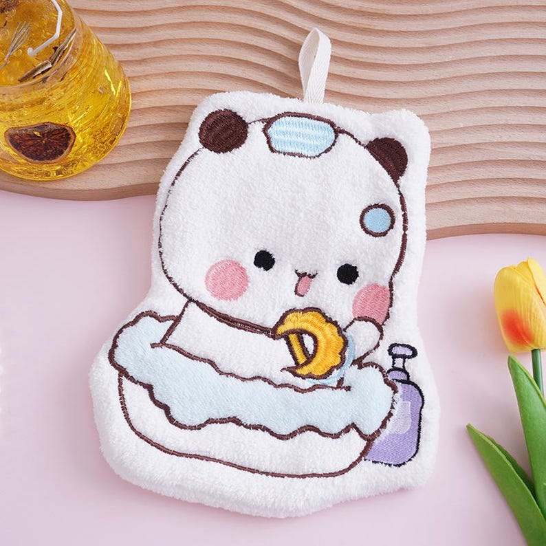 May include: A white panda-shaped washcloth with brown ears and a blue and white striped headband. The panda is holding a yellow cookie and a purple bottle. The washcloth has a loop for hanging.