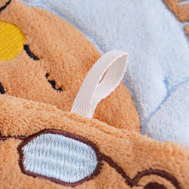 May include: Close-up of a plush, light brown towel with embroidered details. Features a cartoon design with a yellow sun, black eyes, and a light blue oval shape. A white fabric loop is attached to the towel.