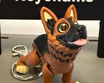 May include: A brown and black articulated German Shepherd dog keychain. The dog has a black nose and tongue sticking out. The keychain is made of plastic and has a silver metal ring.
