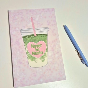 May include: A pink floral notebook featuring a matcha drink design. The illustration shows a clear cup with a pink straw and a heart-shaped label saying "Never too Matcha." A light blue pen is placed on the white surface beside the notebook.