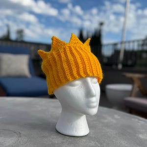 May include: A bright yellow, crocheted crown hat with a spiky design. The hat is displayed on a white mannequin head against a backdrop of a blue sky with clouds. The hat is made of a ribbed knit.