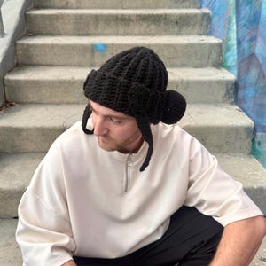 May include: A black, hand-knitted hat with ear flaps and a rounded top. The hat is made of a thick, textured yarn. The person is wearing a cream-colored long-sleeve shirt and black pants.