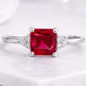 May include: A silver ring featuring a large, square-cut, vibrant red gemstone as the centerpiece. Flanking the central stone are smaller, triangular clusters of white gemstones. The ring is set on a white surface.