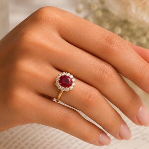 Puede incluir: Un anillo de oro con un rubí rojo intenso de corte ovalado en el centro, rodeado por un halo de pequeños diamantes blancos redondos y claros. La banda del anillo es fina y de oro pulido, mostrando un diseño clásico con una piedra roja vibrante.