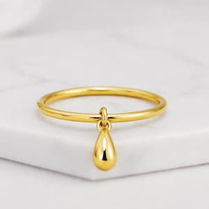 Op de afbeelding: Een goudkleurige ring met een fijne band en een druppelvormige bedel. De gepolijste ring reflecteert licht, en de druppelvormige bedel hangt eronder. De ring staat tegen een witte achtergrond, wat het eenvoudige, elegante ontwerp benadrukt.