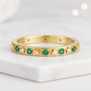 Könnte beinhalten: Ein goldener Ring mit einem abwechselnden Muster aus runden, leuchtend grünen Edelsteinen und kleinen, polierten fünfzackigen Sternen. Das Metall hat eine glatte, reflektierende Oberfläche und die Steine sind sicher in den Ring eingefasst.