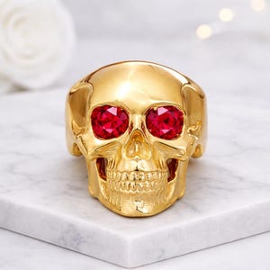 Puede incluir: Un anillo de calavera dorado con grandes ojos de piedras preciosas rojas. El anillo es muy detallado, con dientes y rasgos faciales visibles. El anillo se exhibe sobre una superficie de mármol blanco.