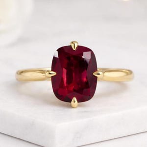 Puede incluir: Un anillo de oro con una gran gema roja oscura con talla cojín. La gema está sujeta por cuatro garras de oro. La banda del anillo es de oro pulido simple. El anillo está sobre una superficie de mármol blanco.