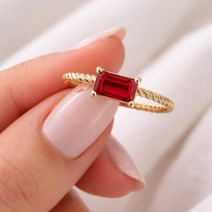 Peut inclure: Une bague en or avec une pierre précieuse rouge foncé de forme rectangulaire. La bande de la bague présente un motif torsadé. La pierre est sertie de quatre petites griffes en or. La bague est sur fond blanc.