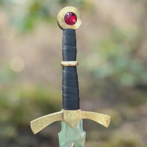May include: A sword hilt featuring a black wrapped grip, a gold-colored crossguard with intricate etched patterns, and a circular pommel topped with a faceted, deep red gemstone. The metal crossguard extends outward with curved, tapered ends.