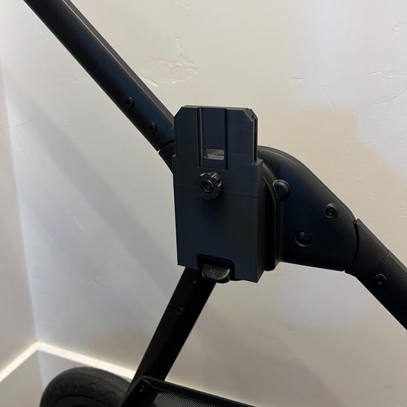 May include: Black stroller attachment with a rectangular mounting bracket and a circular adjustment knob. The attachment is connected to a curved handle and a straight support bar. The stroller wheel is visible in the lower left corner.