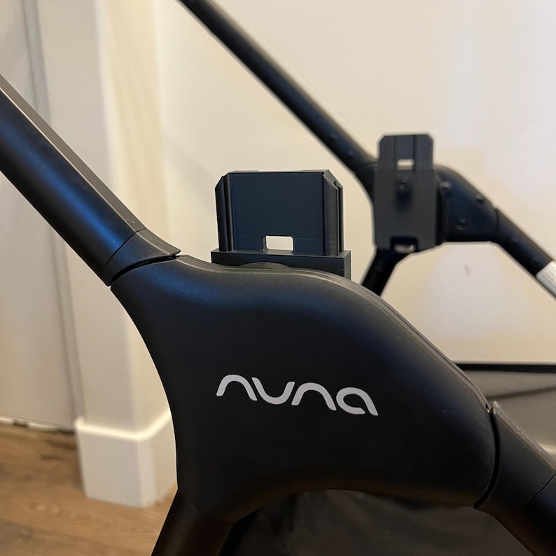May include: Close-up of a black stroller frame with the brand name "nuna" in white lettering. The stroller has a sleek, modern design with a black plastic attachment. The image highlights the stroller's construction and design.