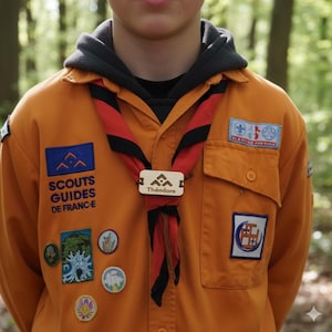 May include: An orange Scouts Guides de France uniform with a red and black neckerchief. The uniform has several patches and a wooden name tag that reads "Théodore". The background is a blurred forest.