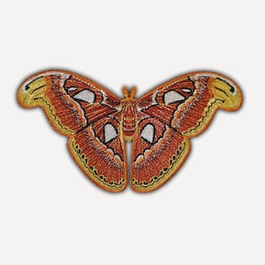Può includere: Una toppa ricamata a forma di falena Atlas con ali nelle tonalità dell'arancione bruciato, rosso intenso e giallo. Le ali presentano motivi complessi con accenti triangolari bianchi, contorni marrone scuro e una finitura in filo testurizzato.