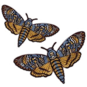 Könnte beinhalten: Zwei gestickte Mottenaufnäher. Die Aufnäher zeigen detaillierte Flügel in Blau-, Gold- und Brauntönen. Der Körper der Motte ist blau und gold gestreift. Die Aufnäher befinden sich auf weißem Hintergrund.