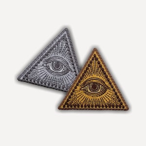 May include: Two triangular embroidered patches featuring an eye centered within a radiating sunburst pattern. One patch is silver and grey, while the other is gold and dark brown. Both patches have a stitched border and a flat, textured surface.