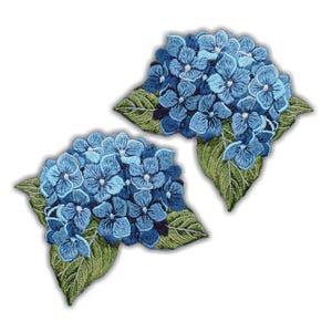 Op de afbeelding: Twee geborduurde patches met blauwe hortensiabloemen met groene bladeren. De patches hebben gedetailleerde stiksels, die de bloemblaadjes en het blad laten zien. De bloemen zijn helderblauw en de bladeren zijn een mix van groentinten.