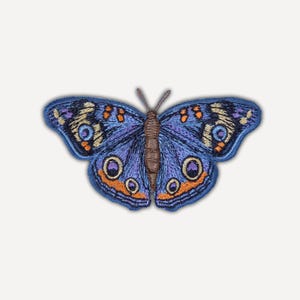 Op de afbeelding: Een geborduurde vlinderpatch met een blauw lichaam en vleugels met ingewikkelde details in tinten paars, oranje en crème. De vleugels vertonen cirkelvormige, oogachtige patronen met donkere centra, omringd door levendige oranje en blauwe randen.