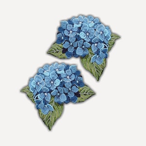 Può includere: Due toppe ricamate raffiguranti grappoli di fiori di ortensia blu con foglie verdi dettagliate. Ogni toppa mostra un intricato lavoro di filo in varie tonalità di blu, creando un motivo floreale strutturato con cuciture visibili.