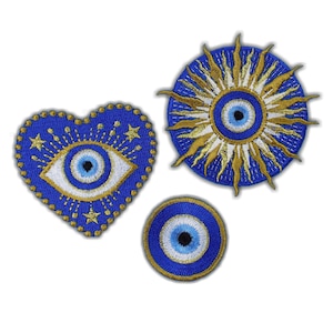 Op de afbeelding: Drie geborduurde patches met het boze oog symbool. Eén is hartvormig, een andere is cirkelvormig met zonnestralen, en de derde is een kleine cirkel. Allemaal in blauw, wit en goud.