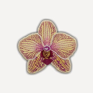 May include: An embroidered patch in the shape of an orchid flower. The petals are a pale yellow color with intricate magenta veins radiating from the center. The central part of the flower features deep maroon and dark brown thread detailing.