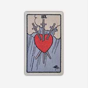 May include: A rectangular embroidered patch featuring the Three of Swords tarot card design. A large red heart is pierced by three grey swords against a light blue background with beige clouds and rain. The Roman numeral III is printed at the top.