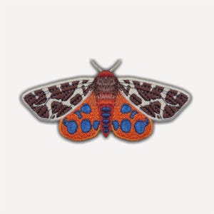 May include: An embroidered patch shaped like a moth with brown and white patterned upper wings. The lower wings are orange with bright blue circular spots. The central body is dark brown with a vertical row of blue accents on the abdomen.