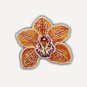 May include: An embroidered patch shaped like an orchid flower with orange and yellow petals, featuring intricate dark red and white details in the center. The edges of the flower are defined by a light-colored stitched border on a flat surface.