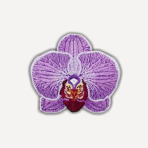 Può includere: Una toppa ricamata a forma di fiore di orchidea. I petali sono di varie tonalità di viola con cuciture bianche lungo i bordi. Il centro presenta dettagli intricati in filo rosso scuro, giallo e bianco, creando un motivo floreale testurizzato.